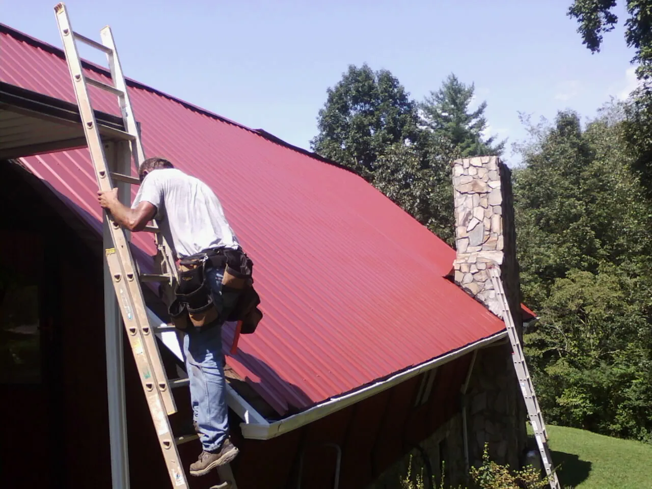 Roofer on a ladder beside a red metal roof and stone chimney — Soffit & Fascia Repair by Roofing Brothers in Blackshear